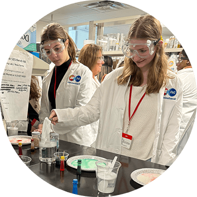 Two young women wear lab goggles and white lab coats. They stand over a black table and hold a white cloth over a beaker filled with liquid. Paper plates holding colored liquids sit on the counter.