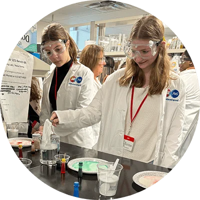 Two young women wear lab goggles and white lab coats. They stand over a black table and hold a white cloth over a beaker filled with liquid. Paper plates holding colored liquids sit on the counter.