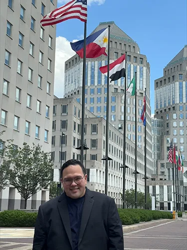 A man stands smiling before a backdrop of tall buildings and a row of international flags.
