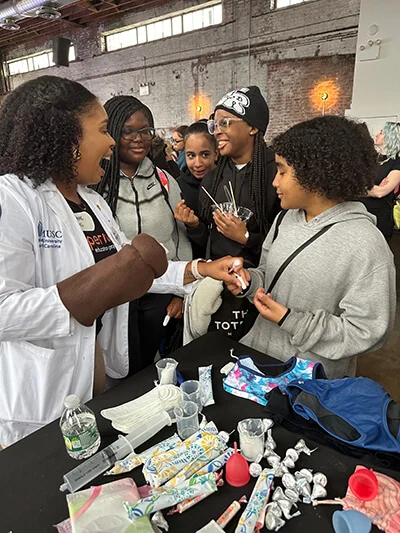 Five young black women gather together. They are smiling at each other as they interact with various period products on a nearby table.