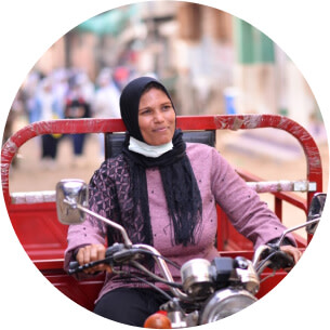 A woman in a pink shirt and black scarf wrapped around her head is driving a moped with a red trailer attached to it.