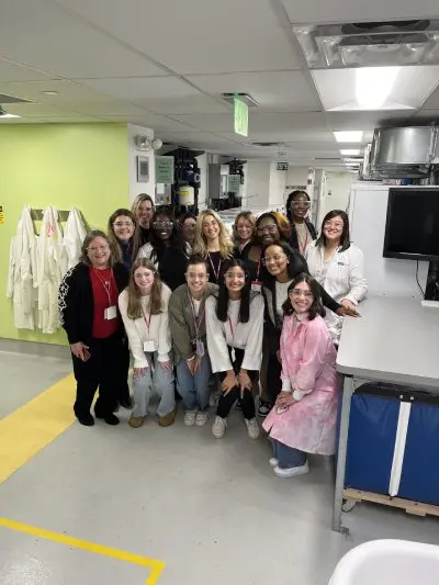 A multicultural group of women and girls pose together. They're wearing lab goggles as they stand together and smile in a laboratory. To their left is a table with a large back screen. Lab coats hang on a lime green wall behind them to their right.