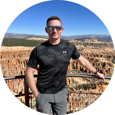 A man in sunglasses and a camouflage shirt stands on a viewpoint, overlooking a vast canyon filled with red-orange rock formations.
