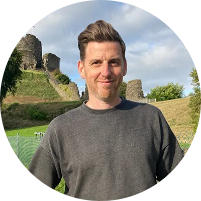 A man with short brown hair wears a grey t-shirt and black pants. He is standing in a field with a small with grass and trees in the background.