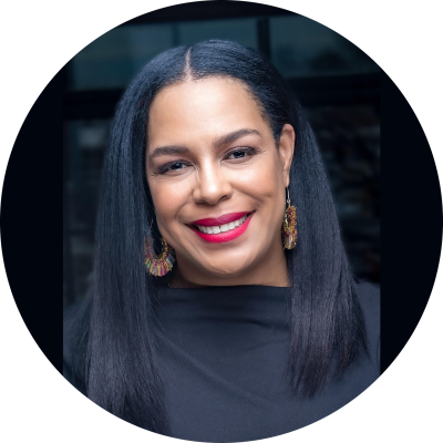 A black woman with long, dark straight hair wears a black blouse and large gold earrings as she smiles at the camera.