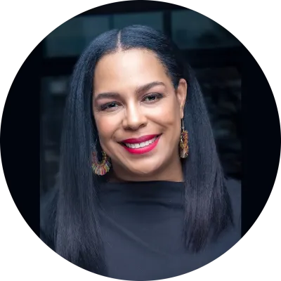 A black woman with long, dark straight hair wears a black blouse and large gold earrings as she smiles at the camera.
