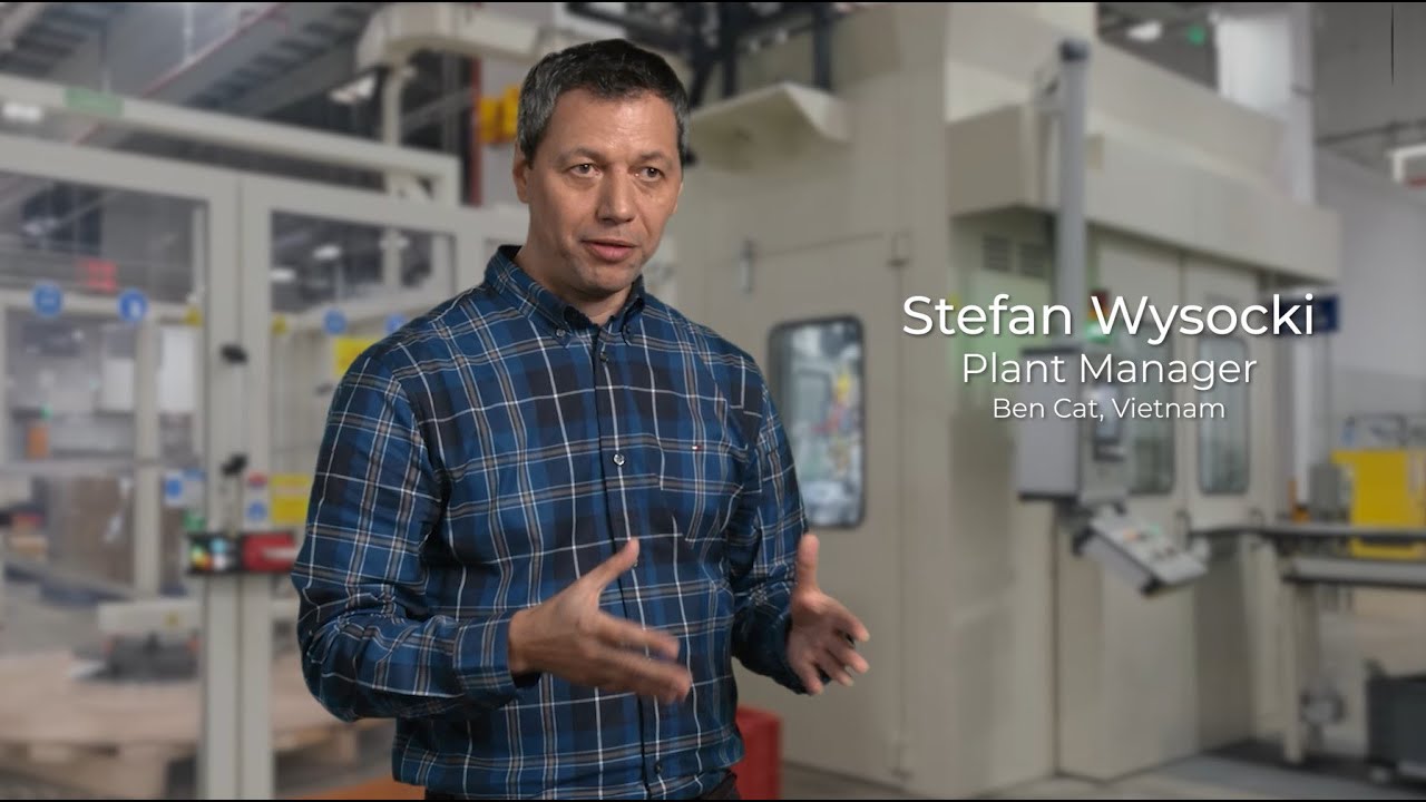 A man in a plaid blue shirt. He stands inside a manufacturing plant.