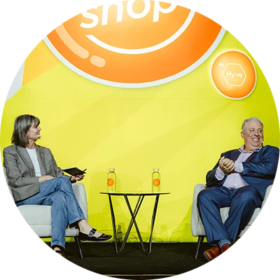A woman and a man sit in armchairs on a stage, engaging in a discussion. The backdrop features a "shop by Hyve" logo and food illustrations.