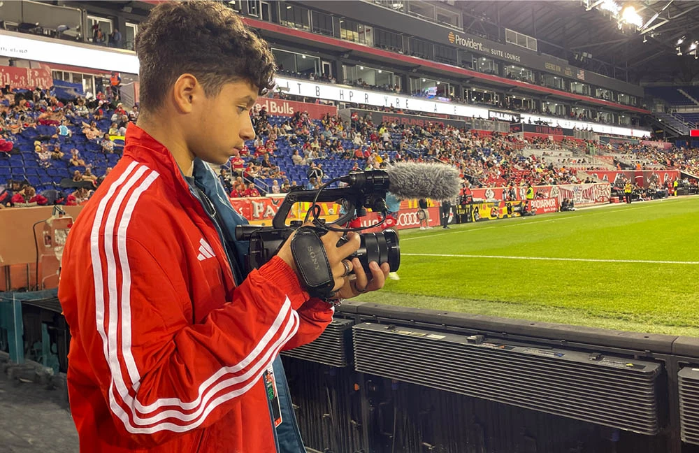 An adolescent Hispanic boy in a red jacket holds a professional camera. He is in the stands of a soccer stadium.