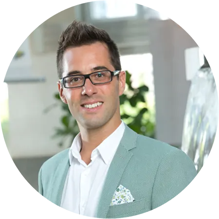 A man with dark hair, glasses, a white shirt and a light green jacket and smiles as he poses for a headshot.
