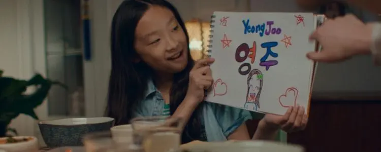 A young Asian girl smiles as she holds up a sign with the American and Korean spellings of her name.