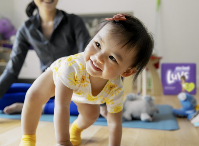 A baby crawls on the floor while their mother looks on from behind.