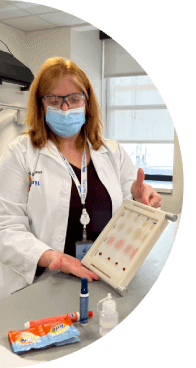 A woman with red hair wearing a white lab coat, mask and safety glasses testing Tide products on stains