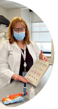 A woman with red hair wearing a white lab coat, mask and safety glasses testing Tide products on stains