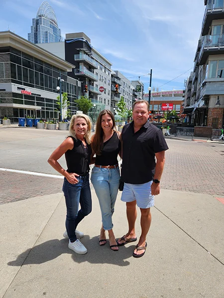 A white woman with short blonde hair, a white woman with long brown hair, and a white man with short brown hair pose together on a sidewalk in Cincinnati. They are all smiling as they face the camera.