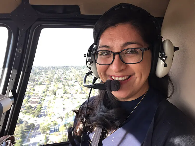 A Hispanic female with long dark hair wears black glasses and smiles from inside the cabin of a helicopter. A city landscape spreads out below in the foreground.