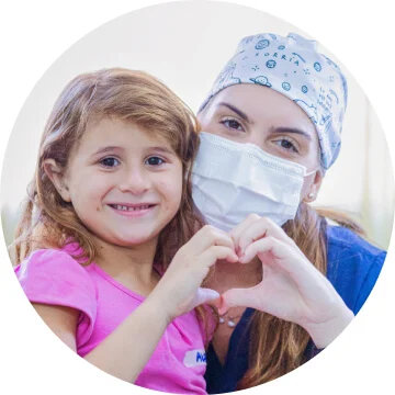 A woman in scrubs and a white medical face mask and a young girl in a pink t-shirt are smiling towards the camera. Their hands are touching to form a heart. 