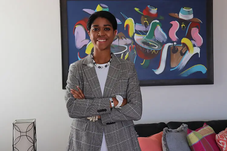 A young black women with dark straight hair, black and white pearl jewelry and a grey and white blazer smiles and poses for a headshot.