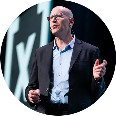 A man in a light blue shirt and black suit jacket. He is wearing a microphone and speaking on a stage.