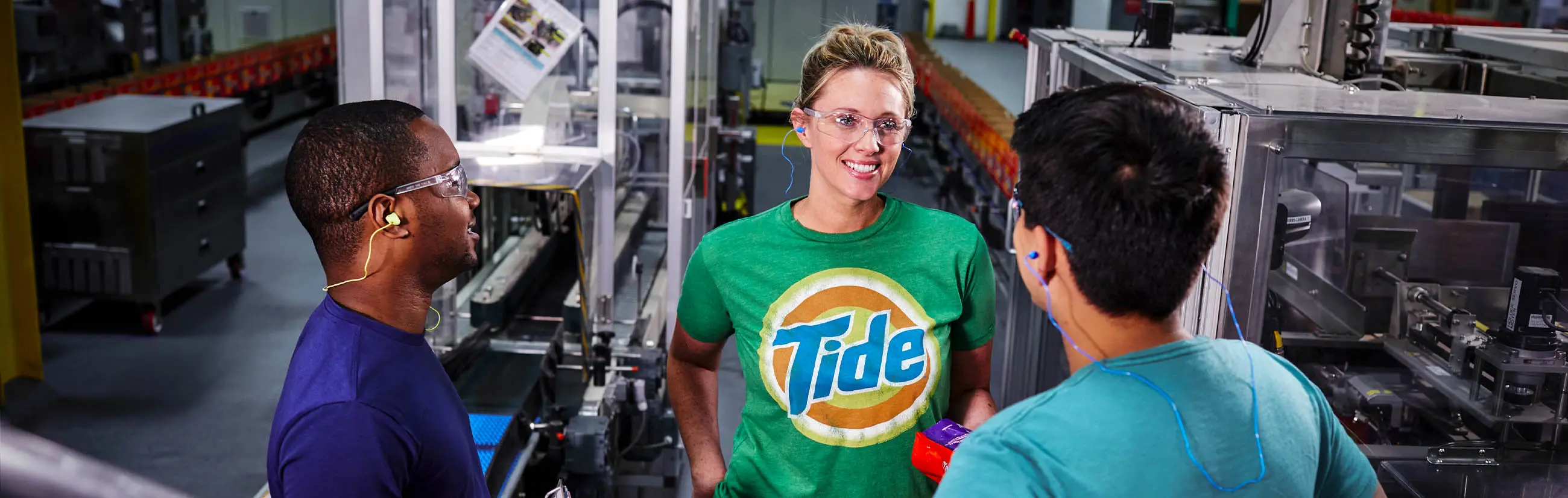 Three P&G plant employees next to a Tide manufacturing line. The female employee facing the camera is wearing a Tide t-shirt.