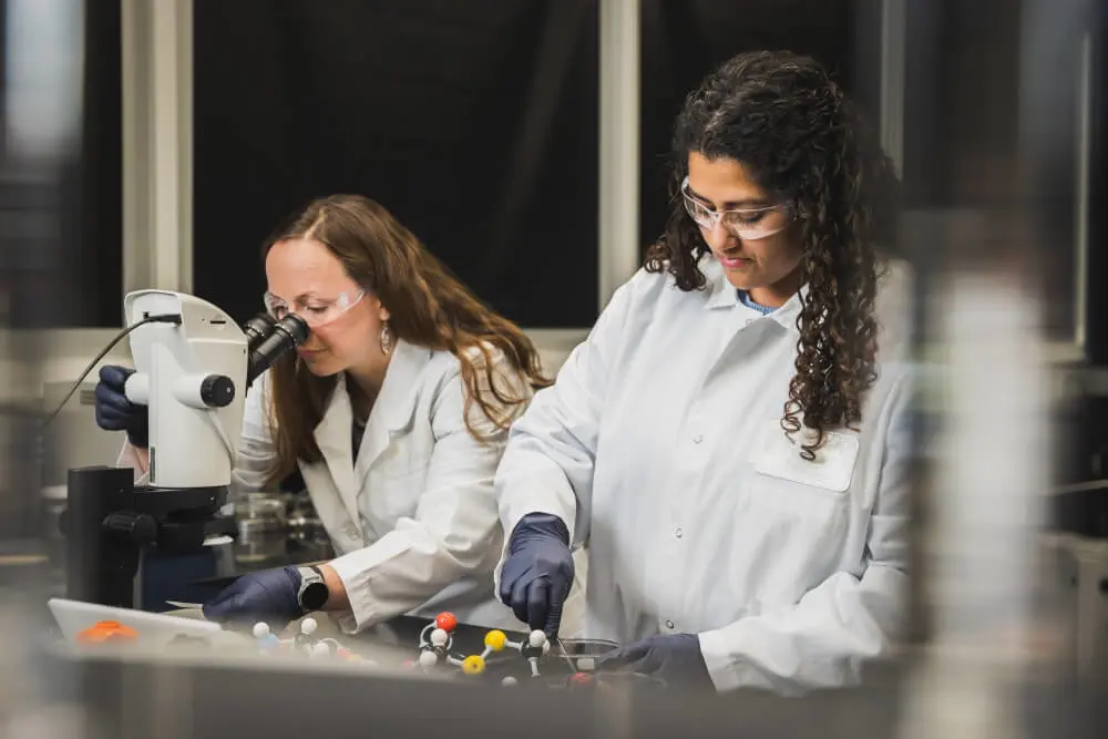 Two women in lab coats and safety glasses work in a laboratory, one looking into a microscope and the other manipulating molecular models.
