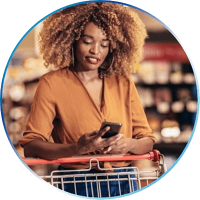 A Black woman with light brown curly hair is wearing an orange blouse. She stands in front of a shopping cart as she looks down at her mobile phone.