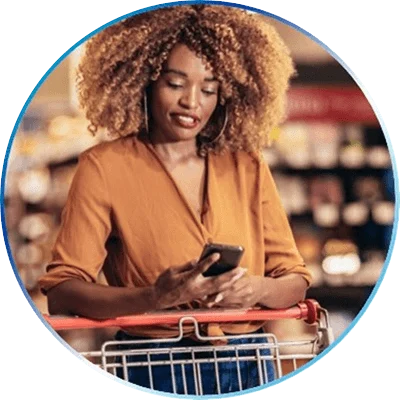 A Black woman with light brown curly hair is wearing an orange blouse. She stands in front of a shopping cart as she looks down at her mobile phone.