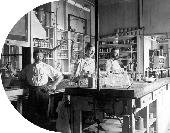 Black-and-white historical photograph of the 1890 first analytical lab at P&G, showing three workers in a laboratory with glassware, bottles, and equipment on wooden benches in an industrial setting.