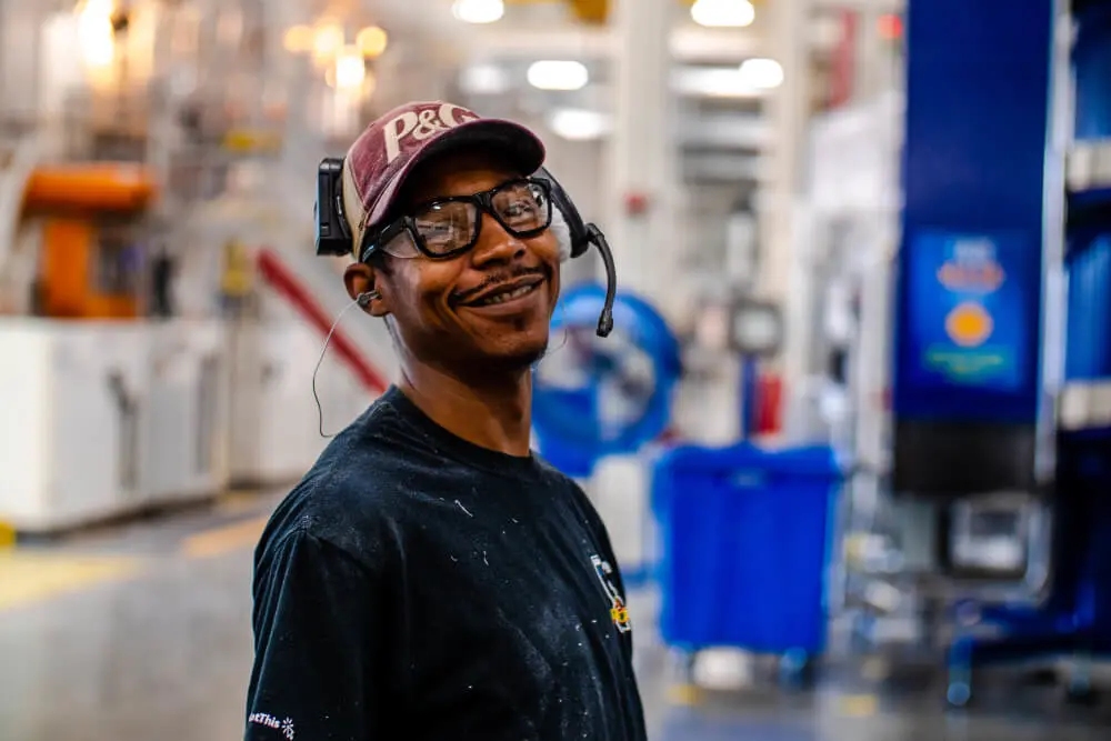 A man in a dark t-shirt, a red P&G baseball cap, safety goggles, earplugs and an audio headset. He smiles, and a manufacturing plant with lots of blue equipment is blurred in the background.