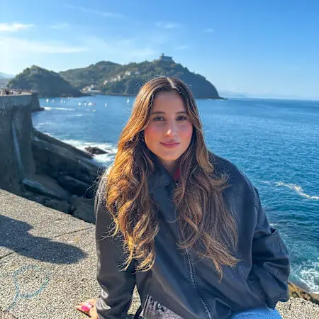 A young woman with long brown hair sits on a cliffside. The ocean and a small island are in the background.