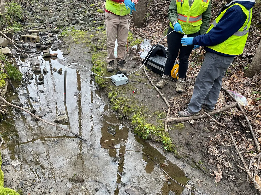  Two individuals in safety gear perform water quality testing by a stream, with tubes extending into the water and equipment on the bank.