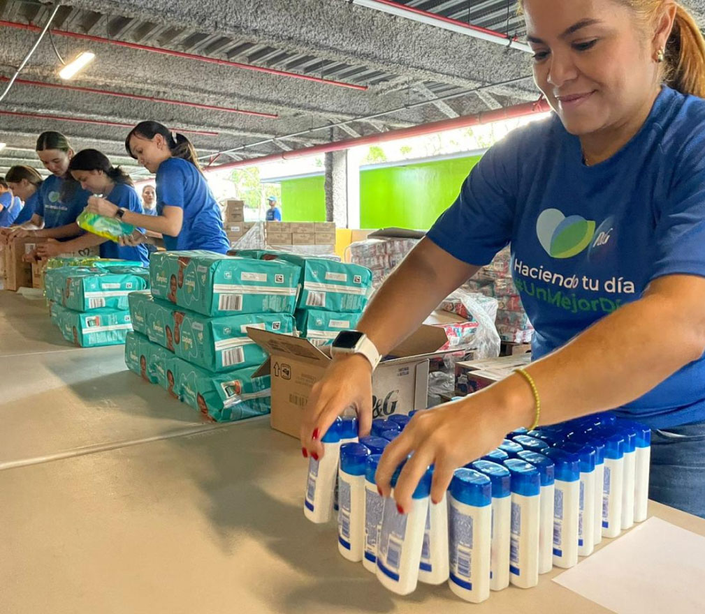 A woman in a blue t-shirt organizing bottles of Head & Shoulders shampoo on a table. There is a pile of Pampers next to her and a group of volunteers further down the table.