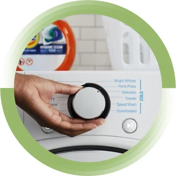 A hand turning the dial of a washing machine. There is a pack of Tide pods and a laundry basket on top of the machine.
