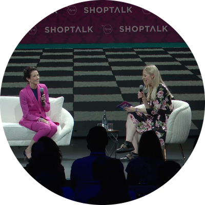 Two women - one in a bright pink pantsuit and the second in a black and pink floral dress - sit together on a dark stage. They each hold a microphone while having a conversation.