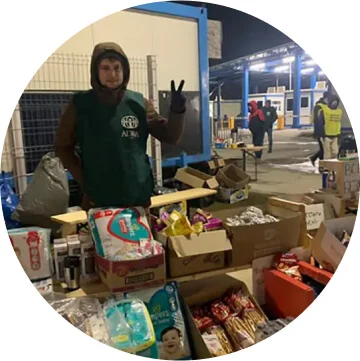 A man in a black sweatshirt is standing behind an array of products like diapers, water and food. His hand is forming a peace sign.