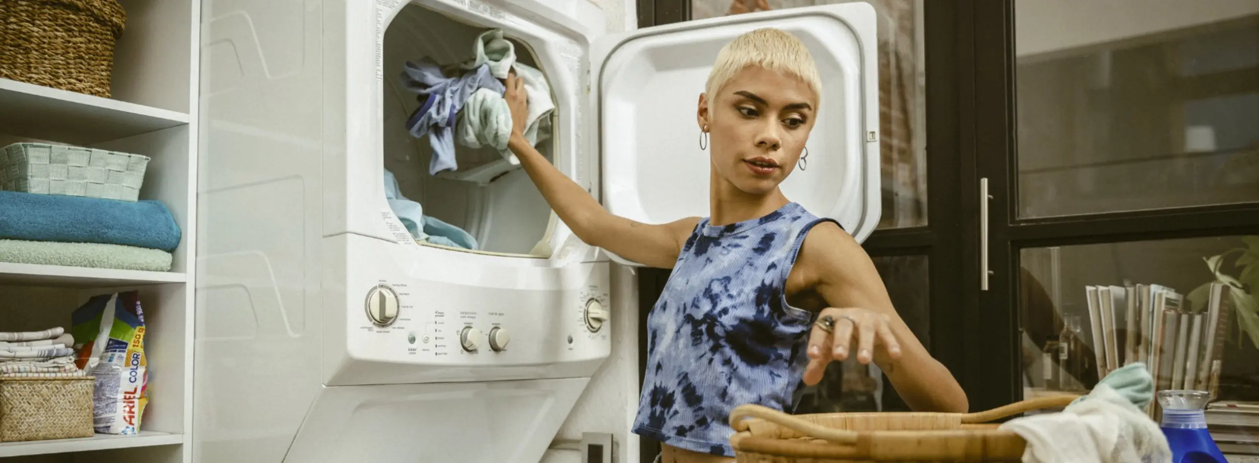 Woman puts laundry into dryer with one hand while reaching into laundry basket with the other.