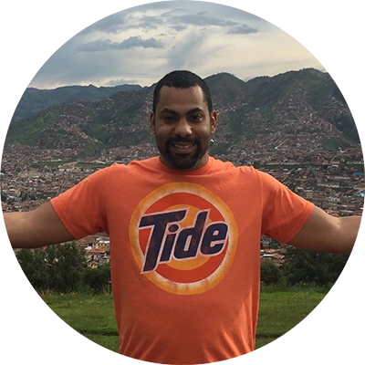 A smiling man in an orange Tide t-shirt opens his arms wide on a green hill overlooking a large valley city and mountains.