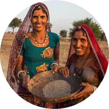Two women in blue and red clothing and read scarves holding a large brown bowl full of Guar. 