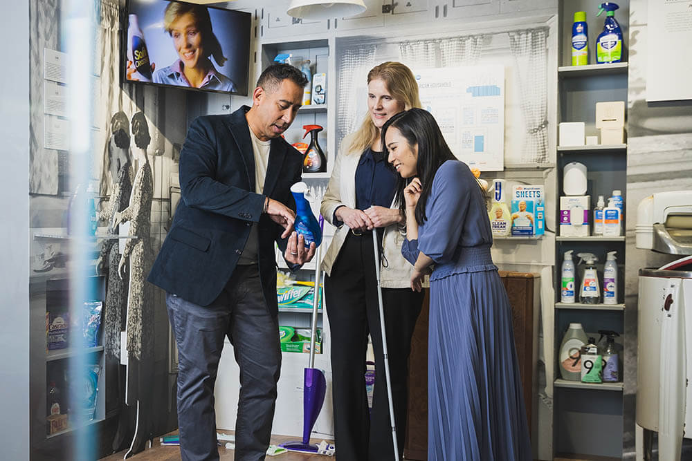 A man holds and points at a bottle of dish soap. Two women stand next to him and lean in to take a closer look.