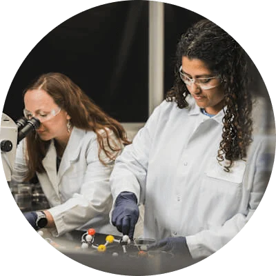 Two women in lab coats are working in a lab. One of them is looking through a microscope. The other is working with a multi-colored molecular model.