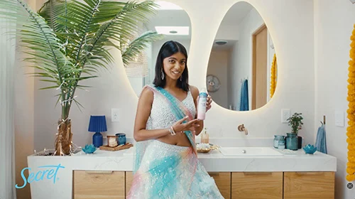 Individual holding a Secret product in a bathroom, with a round mirror, plants, and personal care items on the counter.