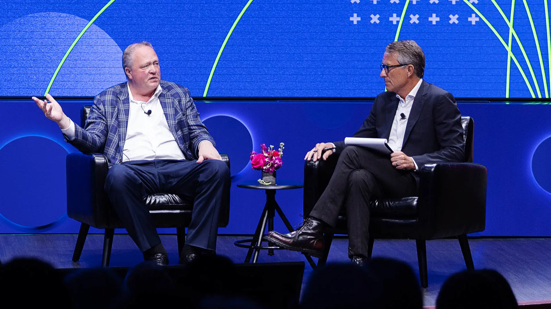 Two white men in business suits sit and talk on a stage. Colorful blue graphics are displayed in the background.