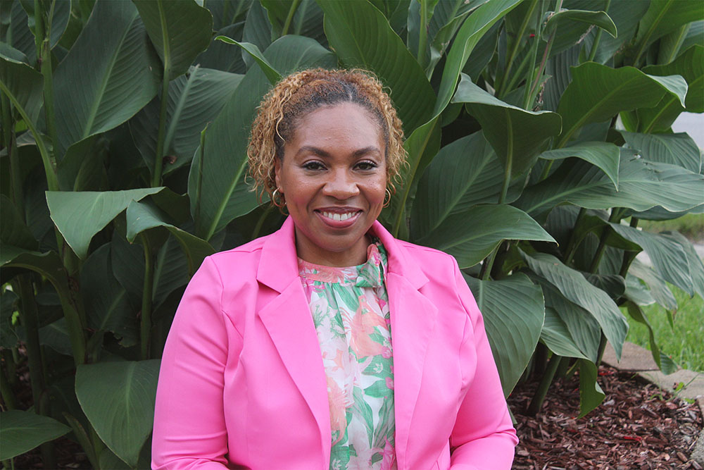 A black women with dark and light colored hair smiles at the camera. She is wearing gold hoop earrings, a pink and green floral patterned blouse and a bright pink jacket.