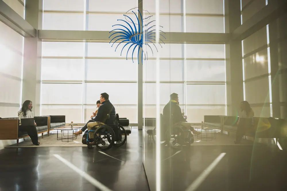 Three P&G employees, one of which is in a wheelchair, sit before a wall of windows in one of P&G's meeting spaces.