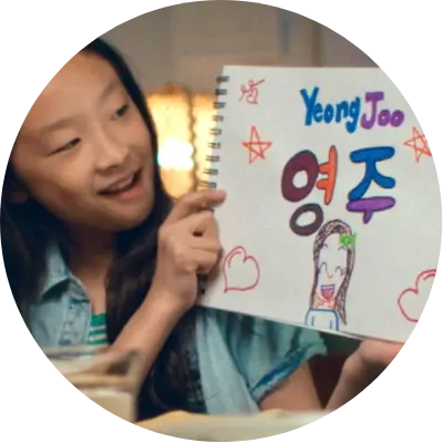 A girl smiles and holds up a drawing of herself and her name "Yeong Joo"