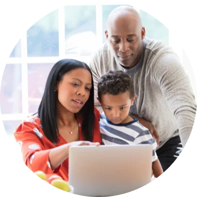 Two young black parents and their young son sit at their kitchen table and look at a silver laptop.