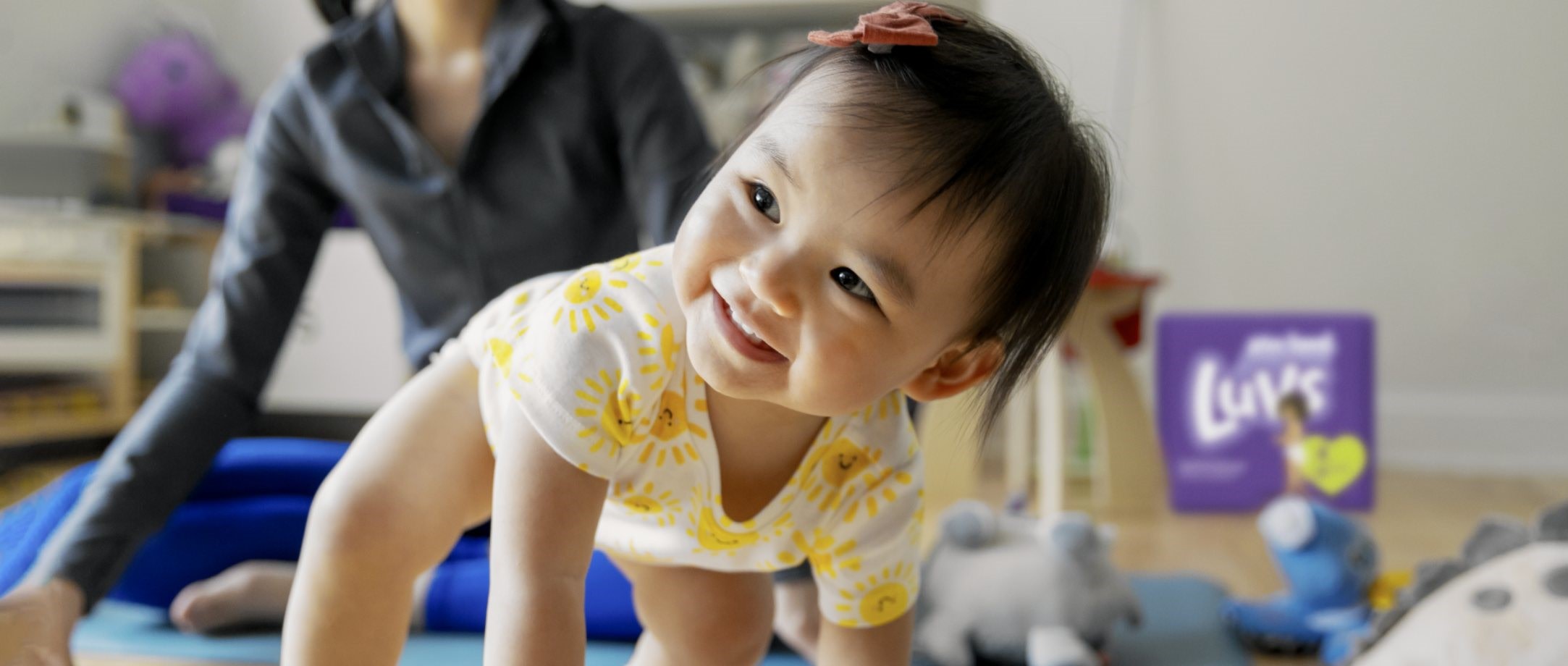 A baby crawls on the floor while their mother looks on from behind.