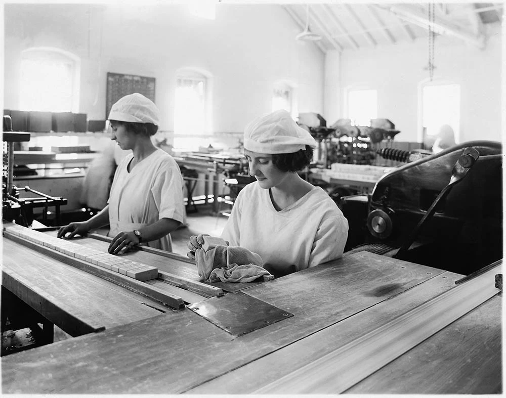 Two young women in white caps and uniforms. They stand in a manufacturing facility behind a long table with a long row of soap bars. The photo is from 1910.