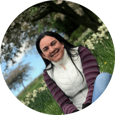Woman sitting on the ground in a field in front of yellow lilies and large trees. She’s wearing jeans, a long sleeve shirt and white vest.