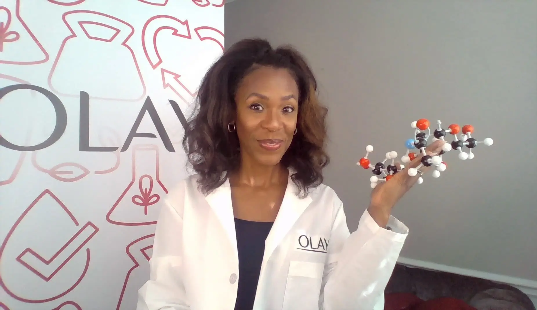 A Black woman with shoulder length hair stands in a labcoat that reads Olay holds a molecular structure with Olay signage in the background.
